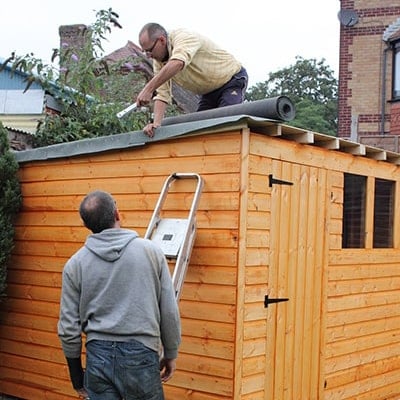 man on top of wooden shed applying felt with another man in the garden watching