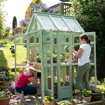 2 women tending to plants in a small, green greenhouse