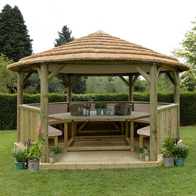 A wooden gazebo with a thatched roof, furnished with benches and a table