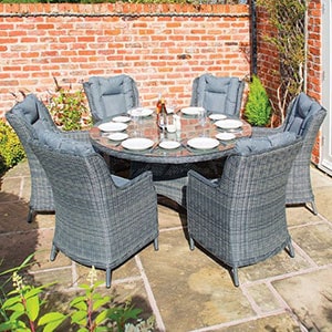 A grey garden furniture set, including 6 rattan dining chairs and a round, glass-top table, all situated on a patio.